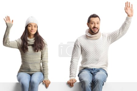 Foto de Joven hombre y mujer con jerséis de invierno sentados en un panel en blanco y saludando aislados sobre fondo blanco - Imagen libre de derechos