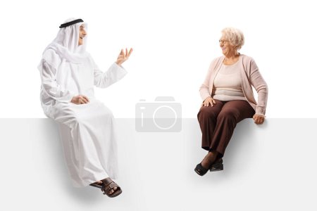 Foto de Hombre árabe con bata étnica y una mujer mayor sentada en un panel en blanco y hablando aislado sobre fondo blanco - Imagen libre de derechos