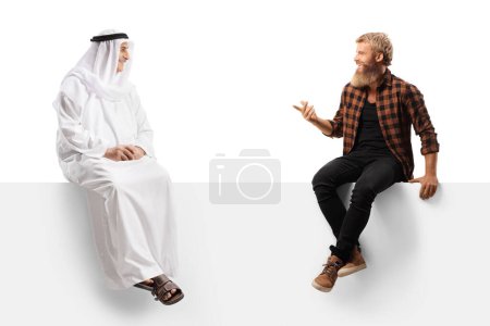 Téléchargez les photos : Jeune homme barbu assis sur un panneau blanc et parlant à un homme arabe isolé sur fond blanc - en image libre de droit