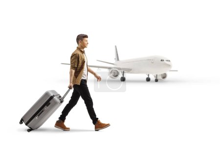 Foto de Foto de perfil completo de un joven pasajero caminando y tirando de una maleta frente a un avión aislado sobre fondo blanco - Imagen libre de derechos