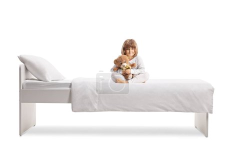 Foto de Niña en pijama abrazando a un osito de peluche y sentada en una cama aislada sobre fondo blanco - Imagen libre de derechos