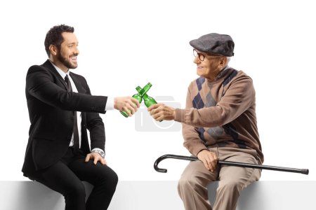 Photo for Elderly man and a young businessman sitting on a blank panel and toasting with beer bottles isolated on white background - Royalty Free Image