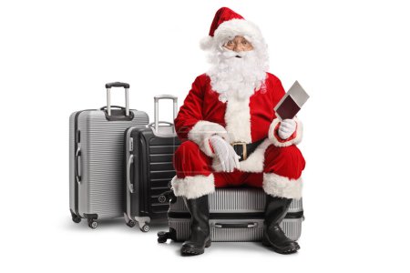 Téléchargez les photos : Père Noël assis sur une valise et tenant un passeport isolé sur fond blanc - en image libre de droit