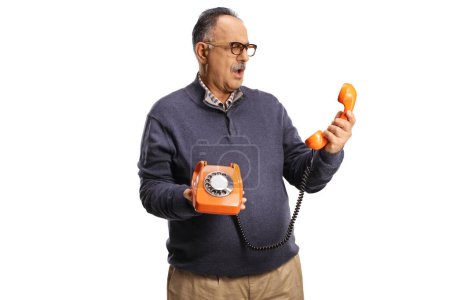 Photo for Confused mature man holding a vintage rotary phone and looking at the handset isolated on white background - Royalty Free Image