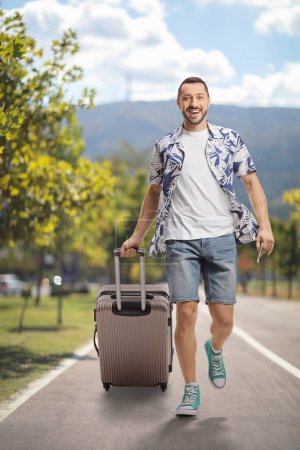 Foto de Retrato completo de un hombre sonriente caminando y tirando de una maleta a la par - Imagen libre de derechos