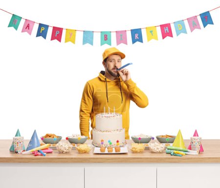 Téléchargez les photos : Casual jeune homme soufflant une corne de fête derrière un comptoir avec du gâteau et favorise isolé sur fond blanc - en image libre de droit