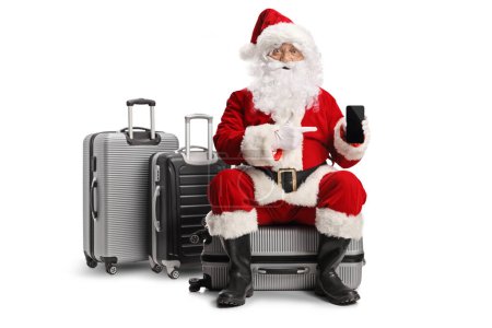 Photo for Excited santa claus sitting on a suitcase and pointing at a smartphone isolated on white background - Royalty Free Image