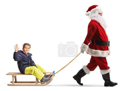 Téléchargez les photos : Plan de profil complet du Père Noël tirant un garçon sur un traîneau en bois isolé sur fond blanc - en image libre de droit