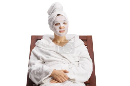 Foto de Mujer joven en un albornoz relajante en una silla de salón con una mascarilla de sábana aislada sobre fondo blanco - Imagen libre de derechos
