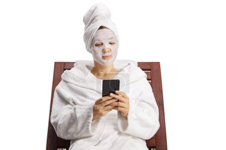 Téléchargez les photos : Femme en peignoir et cheveux enveloppés dans une serviette sur une chaise longue avec un masque facial en drap à l'aide d'un smartphone isolé sur fond blanc - en image libre de droit