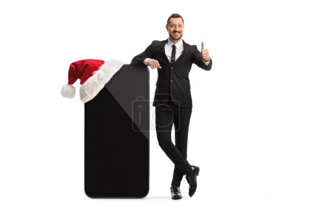 Photo for Businessman standing next to a big mobile phone with a christmas hat and showing thumbs up isolated on white background - Royalty Free Image