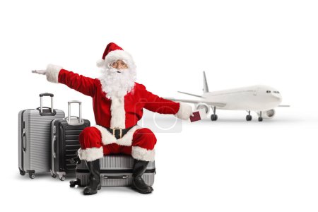 Téléchargez les photos : Père Noël assis sur une valise devant un avion et volant avec les mains isolées sur fond blanc - en image libre de droit