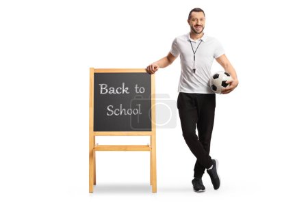 Téléchargez les photos : Professeur d'éducation physique tenant un ballon de football et s'appuyant sur un tableau noir avec texte de retour à l'école isolé sur fond blanc - en image libre de droit