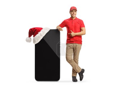 Photo for Full length portrait of a delivery guy pointing at a big mobile phone with a santa claus hat isolated on white backgroun - Royalty Free Image