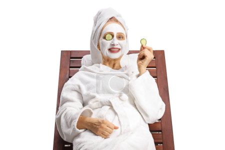 Téléchargez les photos : Femme en peignoir et cheveux enveloppés dans une serviette avec un masque facial et concombre sur les yeux isolés sur fond blanc - en image libre de droit