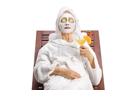 Téléchargez les photos : Femme en peignoir avec masque facial et concombre sur les yeux relaxant avec un cocktail isolé sur fond blanc - en image libre de droit