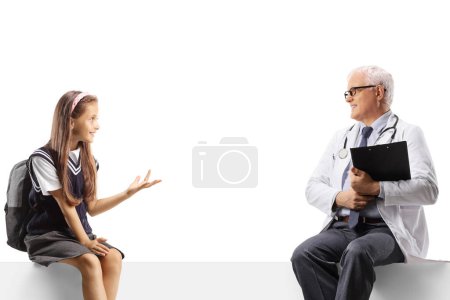 Photo for Schoolgirl sitting on a blank panel and talking to a male doctor isolated on white background - Royalty Free Image