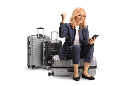 Téléchargez les photos : Femme assise sur une valise regardant un smartphone et gesticulant bonheur isolé sur fond blanc - en image libre de droit