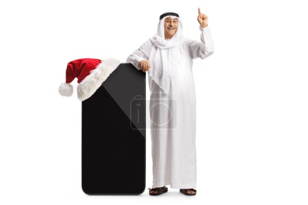 Téléchargez les photos : Homme arabe mature en robe traditionnelle appuyé sur un grand téléphone portable avec un chapeau de Noël et pointant isolé sur fond blanc - en image libre de droit
