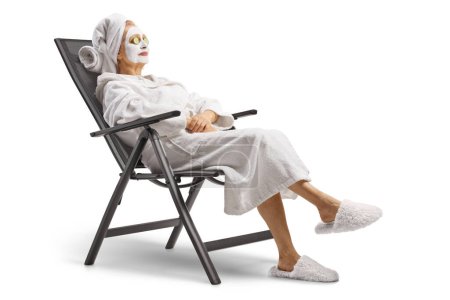 Téléchargez les photos : Femme en peignoir avec un masque facial assis sur une chaise avec du concombre sur les yeux isolés sur fond blanc - en image libre de droit