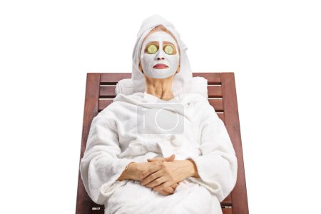 Téléchargez les photos : Femme en peignoir avec masque facial posé sur une chaise longue avec concombre sur les yeux isolés sur fond blanc - en image libre de droit