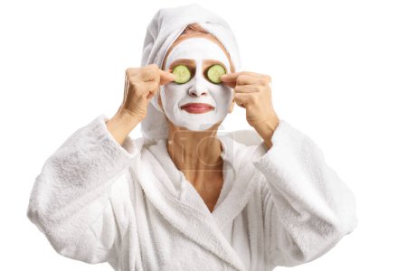 Téléchargez les photos : Femme en peignoir blanc avec un masque facial portant des concombres sur des yeux isolés sur fond blanc - en image libre de droit