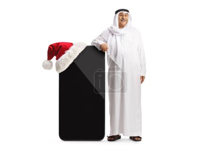 Téléchargez les photos : Arabe en vêtements traditionnels appuyé sur un grand téléphone portable avec santa claus chapeau isolé sur fond blanc - en image libre de droit