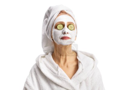 Foto de Mujer en bata de baño con una mascarilla blanca con pepino en los ojos aislados sobre fondo blanco - Imagen libre de derechos