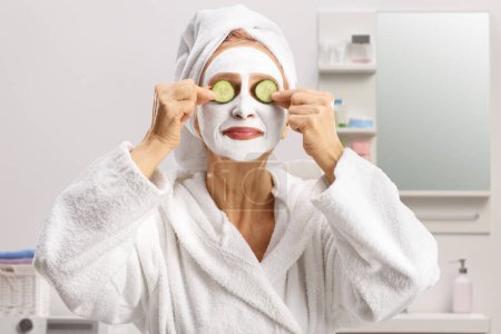 Foto de Mujer en un albornoz con una mascarilla blanca que pone pepino en los ojos dentro de un baño - Imagen libre de derechos