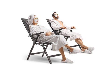Téléchargez les photos : Homme et femme en peignoirs blancs avec des masques faciaux relaxants sur des chaises isolées sur fond blanc - en image libre de droit