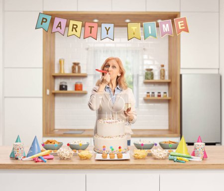 Photo for Casual mature woman blowing a party horn behind a counter with cake and decorations inside a kitchen - Royalty Free Image