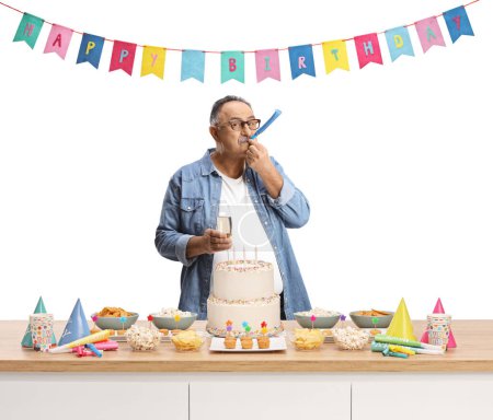 Foto de Casual hombre maduro soplando un cuerno de fiesta de cumpleaños detrás de un mostrador con pastel y decoraciones aisladas sobre fondo blanco - Imagen libre de derechos