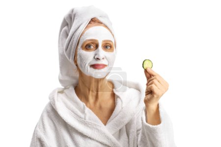 Téléchargez les photos : Belle femme mûre avec une serviette sur la tête et un masque tenant un morceau de concombre isolé sur fond blanc - en image libre de droit