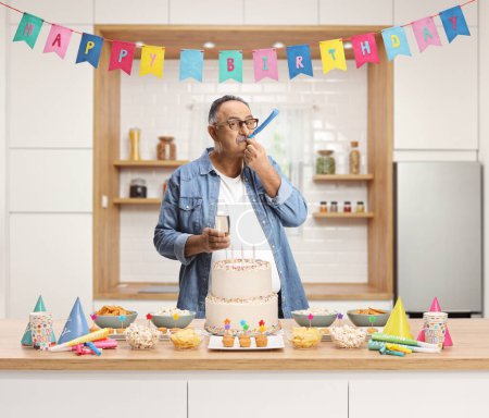 Foto de Hombre maduro soplando un cuerno de fiesta bajo feliz cumpleaños decoración dentro de una cocina - Imagen libre de derechos