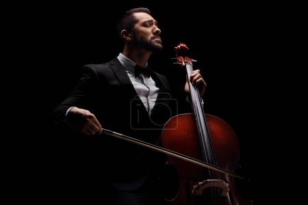 Photo for Male artist in a suit and bow-tie playing a cello isolated over black background - Royalty Free Image