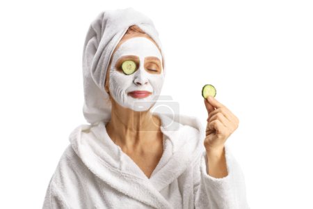 Téléchargez les photos : Femme avec une serviette sur la tête et un masque tenant un morceau de concombre isolé sur fond blanc - en image libre de droit