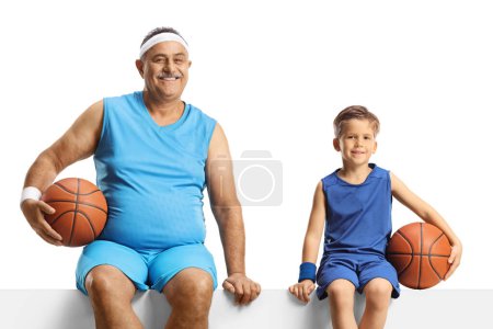 Téléchargez les photos : Homme mûr et un garçon en maillot tenant un ballon de basket et assis sur un panneau blanc isolé sur fond blanc - en image libre de droit