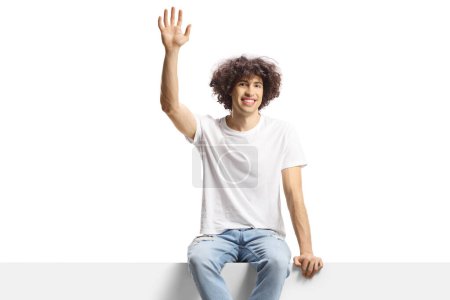 Photo for Casual young man with curly hair sitting on a panel and waving isolated on white background - Royalty Free Image