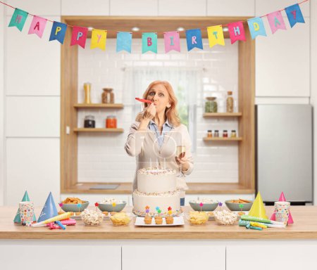 Foto de Mujer con una copa de champán tocando un cuerno de fiesta de cumpleaños detrás de un mostrador con pastel dentro de una cocina - Imagen libre de derechos
