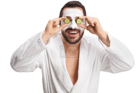 Téléchargez les photos : Jeune homme avec un masque facial tenant le concombre sur les yeux isolés sur fond blanc - en image libre de droit