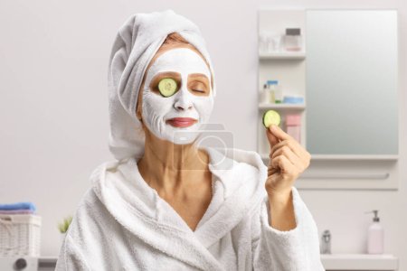 Photo for Mature woman with a towel on her head and a face mask putting cucumber on eyes inside a bathroom - Royalty Free Image