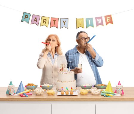 Foto de Hombre y mujer maduros con copas de champán tocando un cuerno de fiesta de cumpleaños detrás de un mostrador con pastel aislado sobre fondo blanco - Imagen libre de derechos