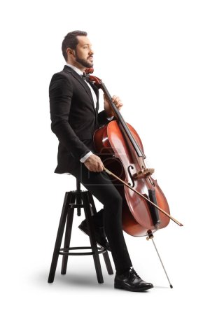 Photo for Young male musician in a black suit and bow-tie sitting on a chair with a cello isolated on white background - Royalty Free Image
