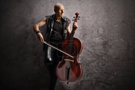 Photo for Punk musician leaning on a gray rugged wall and playing a cello - Royalty Free Image