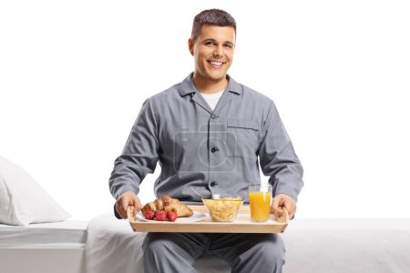 Téléchargez les photos : Jeune homme souriant en pyjama assis sur un lit avec un plateau de petit déjeuner isolé sur fond blanc - en image libre de droit