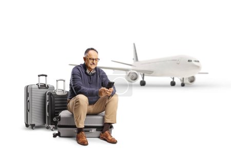Téléchargez les photos : Homme mûr assis sur une valise attendant un vol et utilisant un smartphone isolé sur fond blanc - en image libre de droit