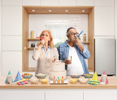 Téléchargez les photos : Homme et femme célébrant et soufflant cornes de fête derrière un comptoir de cuisine - en image libre de droit
