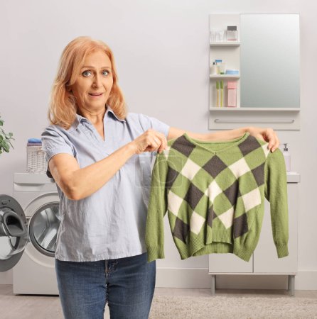 Téléchargez les photos : Femme montrant un pull vert devant une machine à laver dans une salle de bain - en image libre de droit