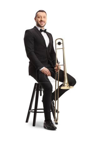 Téléchargez les photos : Musicien masculin en costume noir et nœud papillon assis sur une chaise avec un trombone isolé sur fond blanc - en image libre de droit