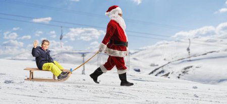 Photo for Full length profile shot of santa claus pulling a boy on a wooden sleigh at a snowy mountain - Royalty Free Image
